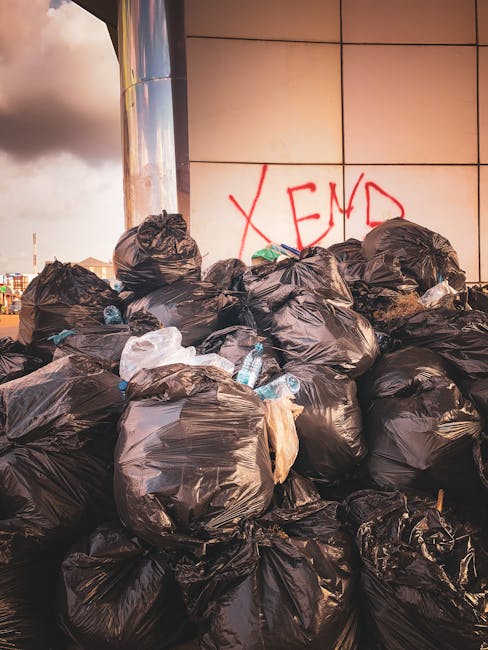 A large collection of tightly tied black plastic rubbish bags stacked against a tiled wall with a glossy finish. Some bags appear bulging with waste, and a few transparent bottles of water and other small debris are visible among the pile. The bags are positioned on what seems to be a pavement or concrete ground, possibly on a street or alleyway. Behind the bags, there's a section of the tiled wall with a graffiti tag in red spray paint spelling 'XEND.' The environment suggests an area where unauthorised or unmanaged waste has been discarded, highlighting the need for professional rubbish removal services like those offered by Rubbish Removal Brent, who specialise in private waste clearance and alternative disposal solutions.