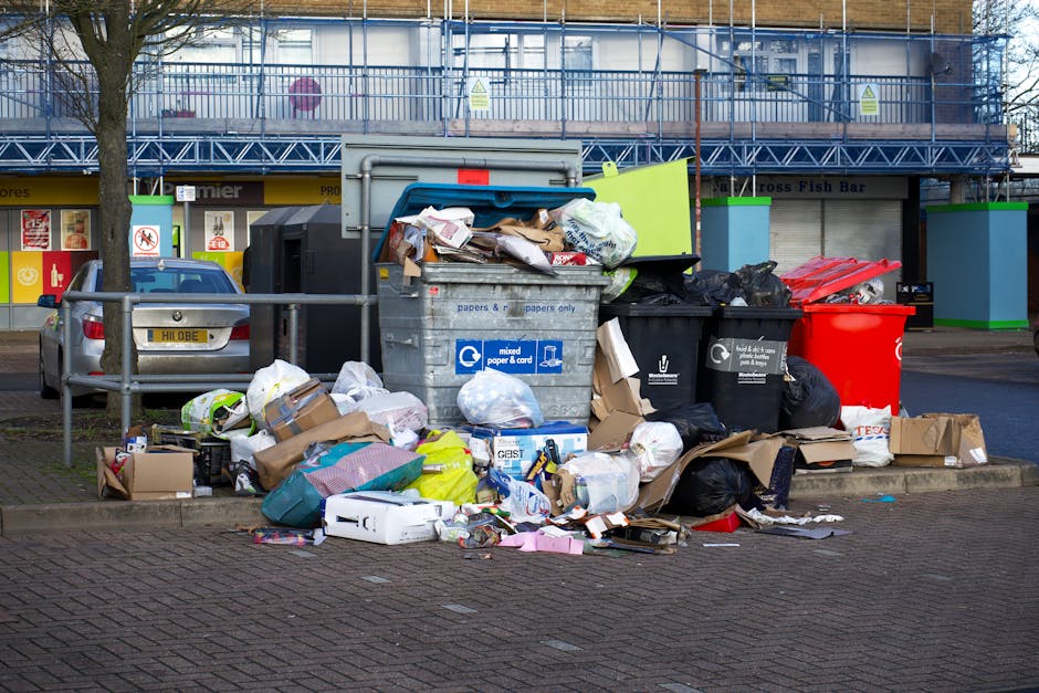 A large collection of rubbish and waste materials piled around a central waste bin on an urban street, with various types of discarded packaging, cardboard boxes, plastic bags, and loose papers spilling onto the paved pavement. The waste bin is grey with a blue lid, designated for mixed paper and cardboard, and is surrounded by additional black rubbish bags and a prominent red recycling bin. Behind the rubbish, a metal railing borders the sidewalk, with a parked silver car nearby. In the background, there is a commercial building wrapped in blue construction scaffolding, with signage for a fish bar and other shops visible at street level. The scene appears to reflect improper disposal or overflow of waste in an area serviced by independent collection, possibly implying a need for professional rubbish removal or on-site clearance, with the environment illuminated by natural daylight, emphasizing the mess and clutter typical of unmanaged rubbish accumulation.