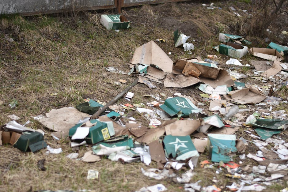 A patch of ground beside a wooden fence shows scattered rubbish, including crushed cardboard boxes, some with visible green and white star logos, and torn paper debris. The cardboard is frayed and weathered, with varying shades of brown and some green print. Small pieces of paper and plastic wrappers are mixed among the larger debris. The ground underneath consists of sparse grass and dirt, with an uneven surface and patches of exposed soil. In the background, the wooden fence has a weathered appearance, with vertical planks and a rough texture. Natural daylight illuminates the scene, casting soft shadows and highlighting the textures of the rubbish and environment, indicating the area is an informal waste dumping spot. This scene exemplifies the typical roadside or vacant lot litter often addressed by private rubbish removal services like Rubbish Removal Brent, emphasizing the need for efficient clearance of disposable waste materials from unmanaged sites.