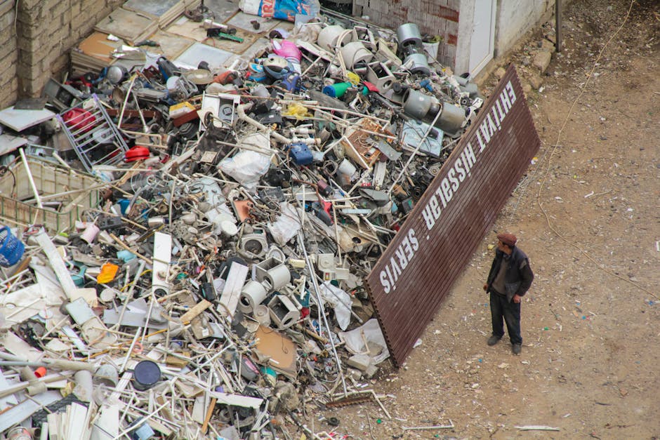 A large pile of mixed household and electronic waste is spread across a gravelly outdoor area beside a brick wall and a white building. The heap contains various materials including broken plastic chairs, metal frames, and damaged electronic appliances such as humidifiers and small appliances, all showing signs of disrepair. The items are scattered and haphazardly piled, with some objects overlapping others. A brown metal barrier with the inscription 'THE FINAL HEADING IN' leans against the waste, separating the debris from the open space. To the right, a man dressed in dark clothing and a brown hat stands observing the waste, surrounded by loose gravel and dirt that form the ground environment. The setting appears to be an industrial or construction site designated for rubbish sorting or disposal, with natural, diffuse lighting illuminating the scene, emphasizing the texture and disorder of the waste materials. This scene reflects the typical environment where private waste collection services may intervene to manage bulky or electronic waste collection efficiently, such as those provided by Rubbish Removal Brent in Brent.