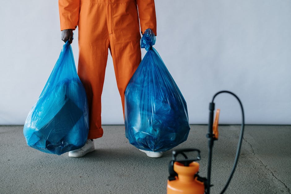 A person dressed in an orange protective suit, with only their lower body visible, is standing on a concrete surface holding two large, blue plastic rubbish bags filled with waste. The bags appear to be tightly packed and slightly transparent, revealing the presence of various material debris inside. The individual is wearing white footwear, and a black glove is partially visible on one hand. In the foreground, there is a partially visible orange and black device, likely a disinfectant or cleaning sprayer, connected to a hose. The background is plain, light-colored, and free of specific environmental features, emphasizing the focus on the waste collection activity. This scene exemplifies a private, on-site waste removal process, representing the kind of service provided by companies like Rubbish Removal Brent for commercial or residential rubbish clearance, particularly suited for contexts such as bulky waste removal from shops or other commercial properties.