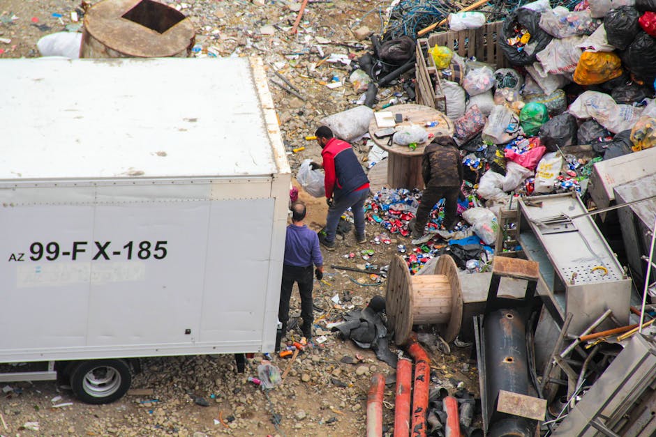 An outdoor scene showing three individuals engaged in rubbish clearance amidst a large accumulation of waste materials. The area is cluttered with numerous black, white, and multicolored plastic bags filled with debris, alongside various discarded items such as wooden reels, metal pipes, and broken appliances. One person, dressed in a red and black jacket, appears to be actively sorting or lifting waste, while another in darker clothing is bending over to pick up items. A third individual, wearing a purple top, stands nearby observing or assisting with the task. To the left, there is a large white truck with a visible registration number '99-FX-185' parked on a gravel surface; the truck's side panel is plain and unadorned. The surrounding environment suggests an unmanaged outdoor waste dumping area, with scattered debris and a mixture of construction and household refuse. This scene illustrates an on-site rubbish removal or waste clearance operation, supporting independent waste disposal activities typical of private rubbish collection services like those offered by Rubbish Removal Brent, especially around areas with accumulated rubbish or during local clean-up efforts.