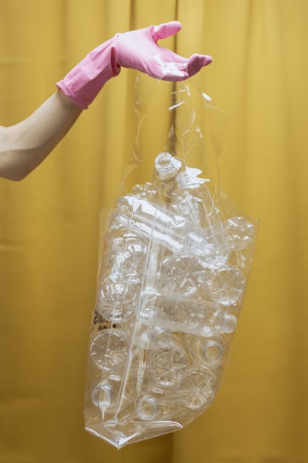 A close-up of a person's hand wearing a pink rubber glove, holding a large, clear plastic bag filled with crumpled and empty transparent plastic bottles, some with visible caps. The background features a solid yellow curtain, providing a neutral backdrop that highlights the recycling activity. The scene documents an aspect of waste management and plastic recycling, which may be related to alternative rubbish collection or private disposal services offered by Rubbish Removal Brent. The plastic bottles are varied in size and shape, with some appearing flattened, indicating they have been previously used and discarded. The bag is held open at the top, ready for collection or further processing, emphasizing the importance of proper waste segregation and recycling efforts. The lighting is bright and even, ensuring the details of the plastic and the gloved hand are clearly visible, aligning with professional waste handling practices and supporting environmentally conscious waste disposal methods.