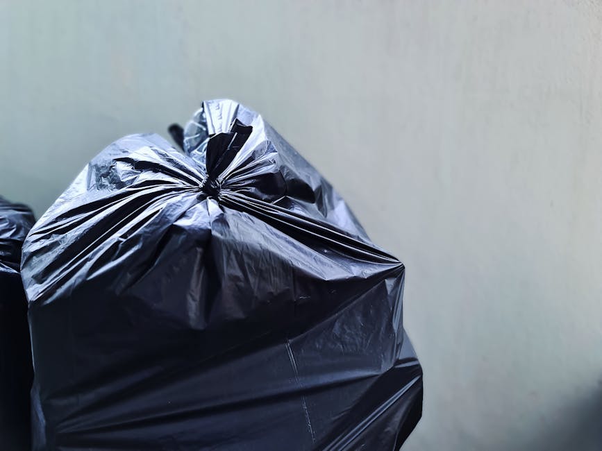 An outdoor scene showing three individuals engaged in rubbish clearance amidst a large accumulation of waste materials. The area is cluttered with numerous black, white, and multicolored plastic bags filled with debris, alongside various discarded items such as wooden reels, metal pipes, and broken appliances. One person, dressed in a red and black jacket, appears to be actively sorting or lifting waste, while another in darker clothing is bending over to pick up items. A third individual, wearing a purple top, stands nearby observing or assisting with the task. To the left, there is a large white truck with a visible registration number '99-FX-185' parked on a gravel surface; the truck's side panel is plain and unadorned. The surrounding environment suggests an unmanaged outdoor waste dumping area, with scattered debris and a mixture of construction and household refuse. This scene illustrates an on-site rubbish removal or waste clearance operation, supporting independent waste disposal activities typical of private rubbish collection services like those offered by Rubbish Removal Brent, especially around areas with accumulated rubbish or during local clean-up efforts.