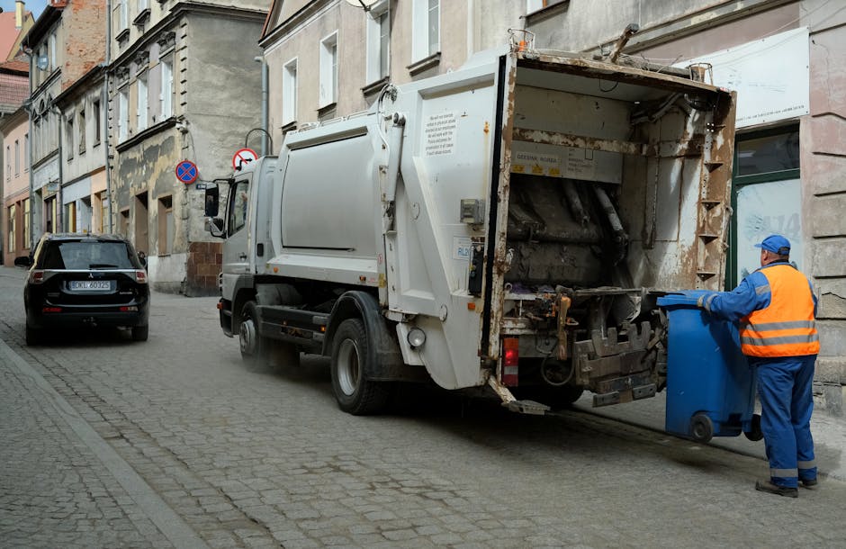 A white industrial rubbish collection truck belonging to Rubbish Removal Brent is parked on a narrow urban street next to a row of multi-storey buildings with beige and grey facades. The truck features a large rear loading hopper, showing signs of rust and wear, with an open waste compaction compartment facing the street. A worker dressed in a blue uniform and an orange high-visibility vest is positioned on the right side of the image, actively tipping a blue wheelie bin into the hopper. The street surface is made of cobblestone paving, with a black car parked on the left side of the road. In the background, there are two traffic signs indicating no stopping and no entry, mounted on a pole attached to one of the buildings. The lighting is neutral, indicating daytime, and the scene captures a typical private rubbish collection process, reflecting the kind of on-site waste handling often carried out by independent waste removal services near Wembley Stadium area.