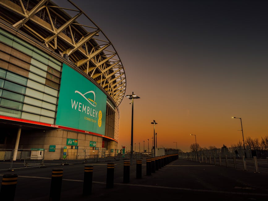 A wide-angle view of Wembley Stadium in Brent, taken during the evening at sunset, features the stadium's distinctive curved roof structure with a metallic finish and a large digital screen displaying the Wembley logo and connecting details. The foreground shows a paved area with low metal barriers, black and yellow-striped bollards, and tall street lamps aligned along the pathway, all set against a gradient sky transitioning from orange near the horizon to darker shades overhead. The scene is well-lit by ambient light, with no visible rubbish or waste materials, but the setting relates to the area where private waste collection or rubbish removal services by Rubbish Removal Brent could be provided for stadium events or surrounding infrastructure.